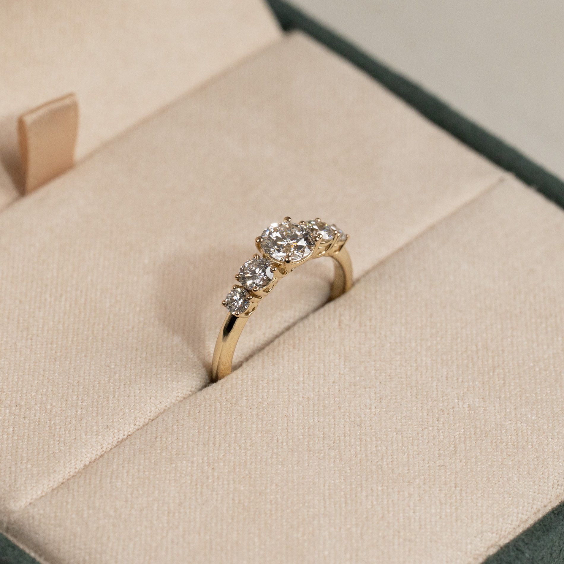 Gold ring with five sparkling round-cut diamonds of graduating size, resting in a beige presentation box.