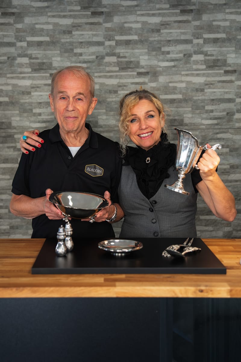 Two smiling people, an older man and a woman, proudly display a collection of silver items.