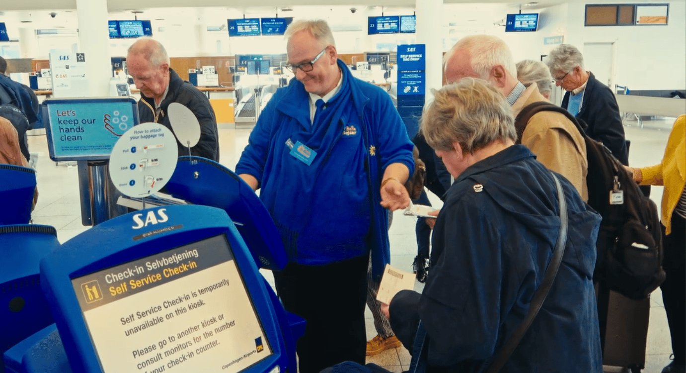Airport staff assists passengers at a self-service check-in kiosk that displays an "unavailable" message.