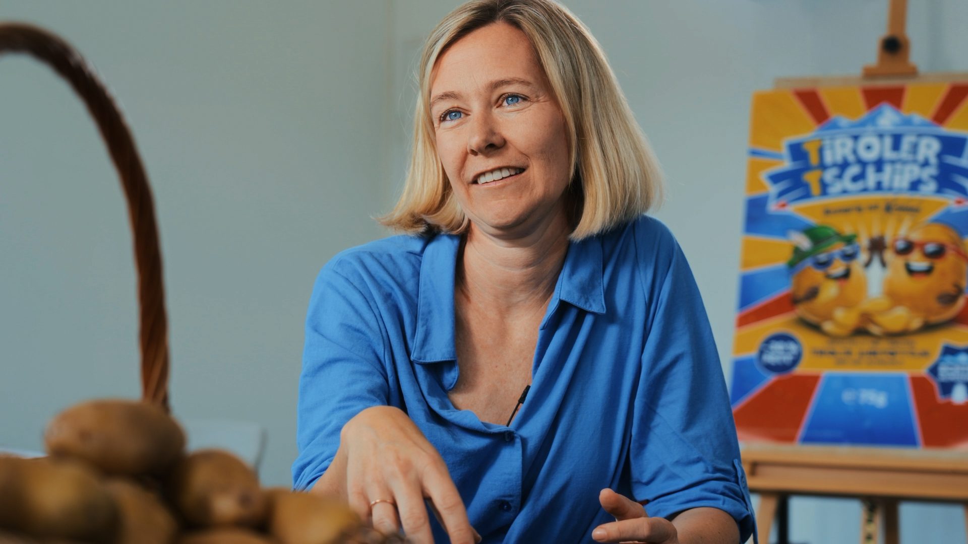 Smiling blonde woman with a basket of potatoes and a "Tiroler Ttschips" sign in the background.