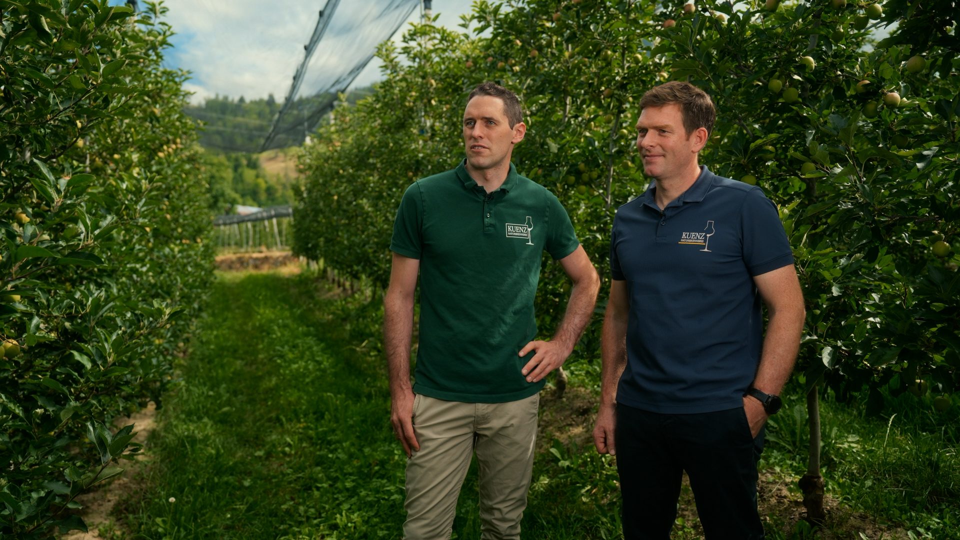 Two men in "KUENZ" branded shirts stand in a lush apple orchard.