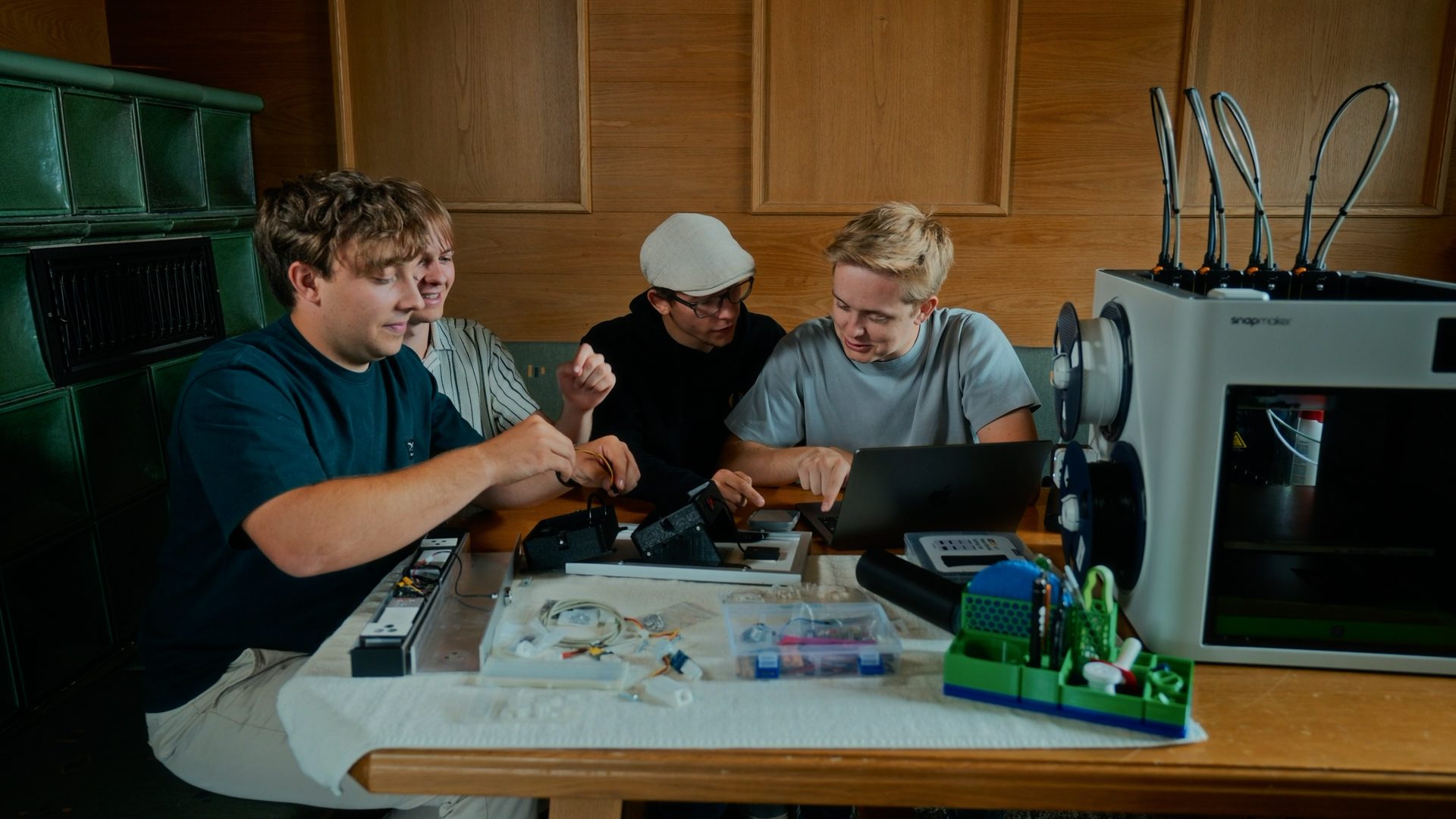 Three young men collaborate on a project, using a laptop and various electronics next to a 3D printer.