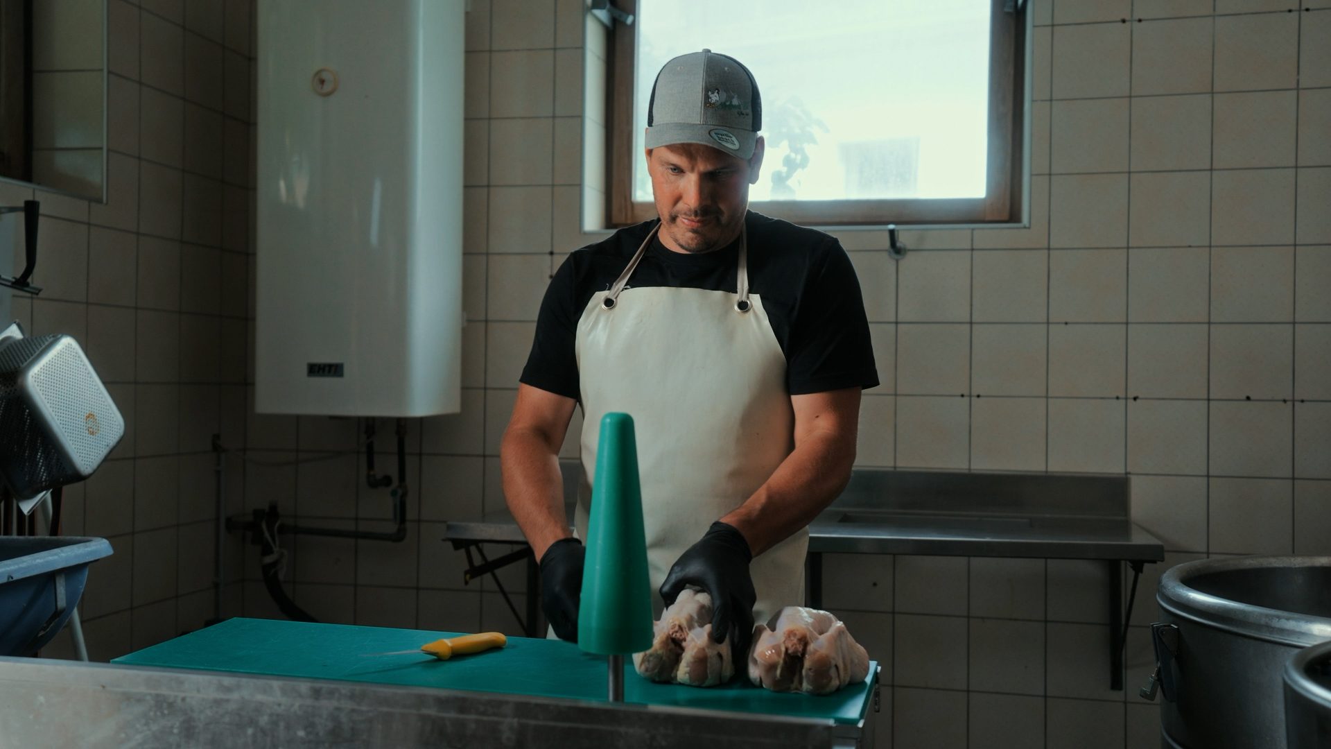 Man in an apron and cap is preparing whole chickens on a green cutting board in a tiled kitchen.