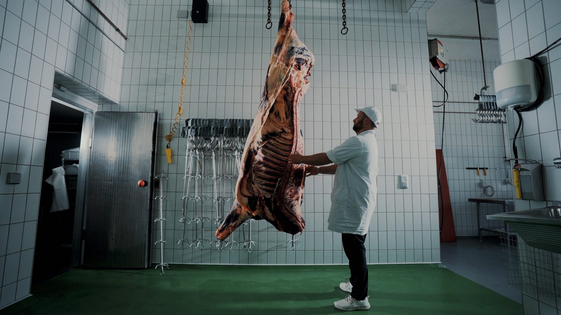Butcher in a white coat inspecting a large beef carcass hanging from a hook in a tiled room.