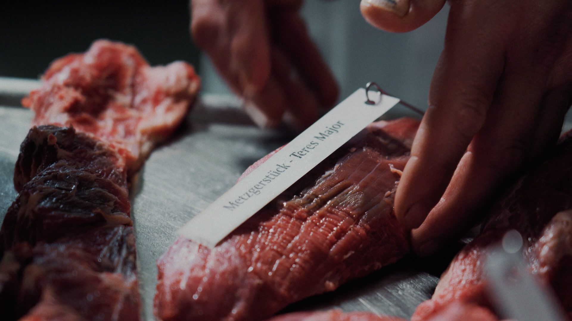 A hand inspects a cut of raw meat labeled "Metzgerstück - Teres Major" on a metallic surface.