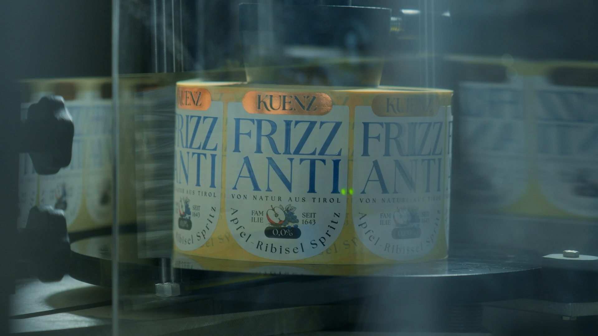 A KUENZ FRIZZ ANTI label, featuring 'VON NATUR AUS TIROL' and '0.0% Apfel-Ribisel Spritz' on a production line.