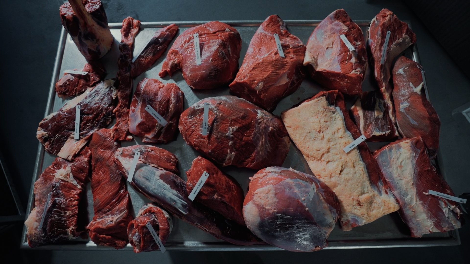 Various raw meat cuts with labels displayed on a metal tray, showcasing butchery preparations.