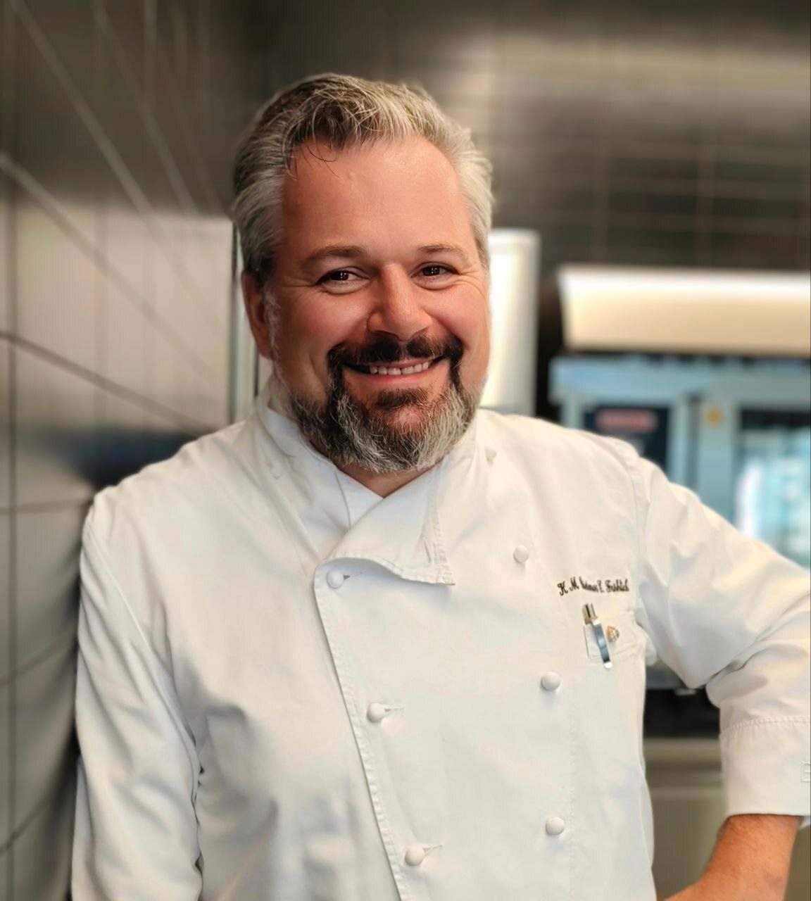 A smiling male chef with graying hair and a beard, wearing a white uniform, leaning in a kitchen.