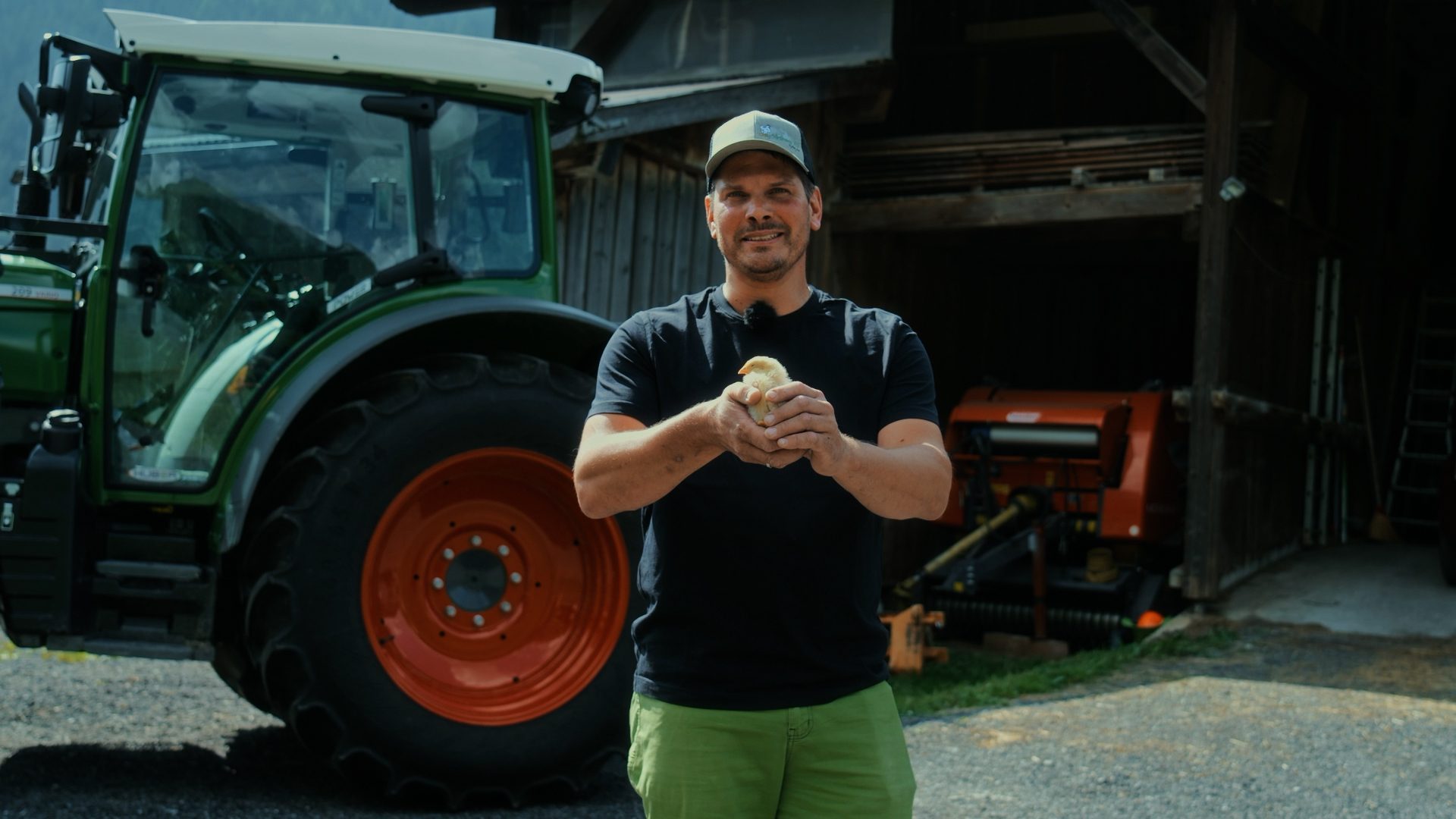 A man in a cap and black t-shirt holds a tiny yellow chick in his hands, standing before a tractor and barn.
