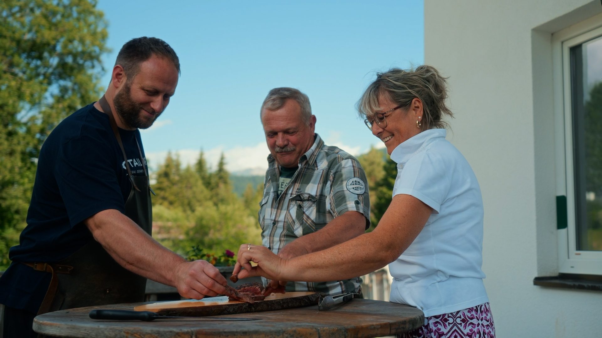 Three people, two men and a woman, smiling and focused on slicing meat outdoors on a wooden table.