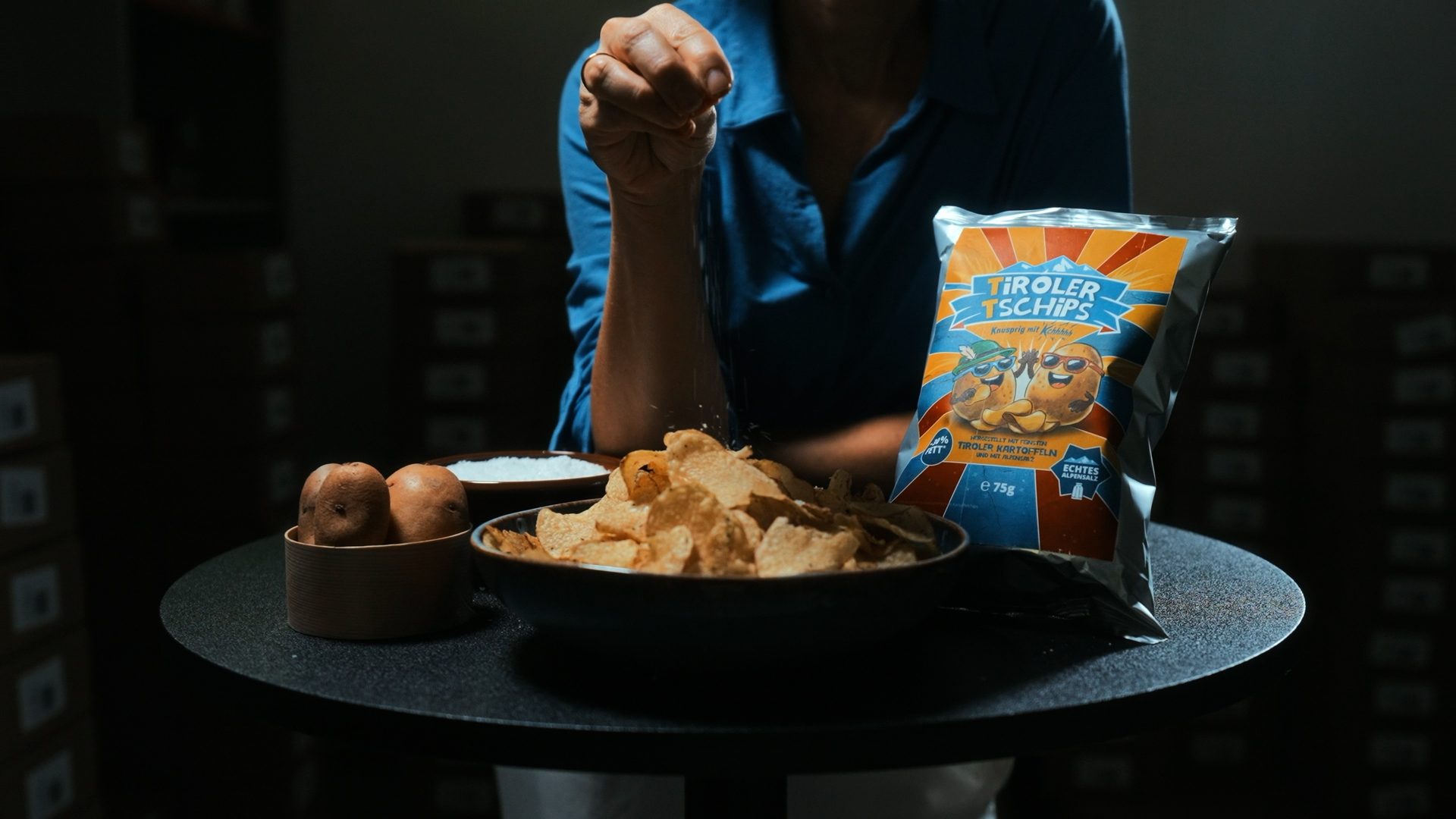 A person sprinkles salt onto a bowl of potato chips next to a bag of "Tiroler Tschips" and raw potatoes.