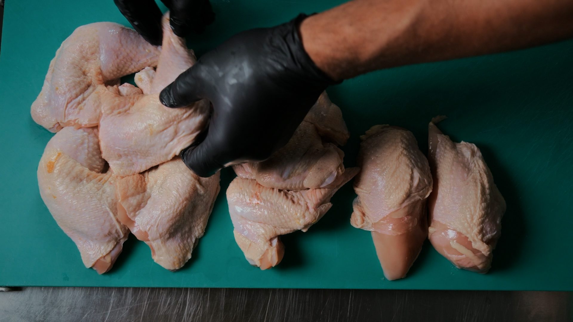 A gloved hand arranges raw chicken pieces on a green cutting board, preparing them for cooking.