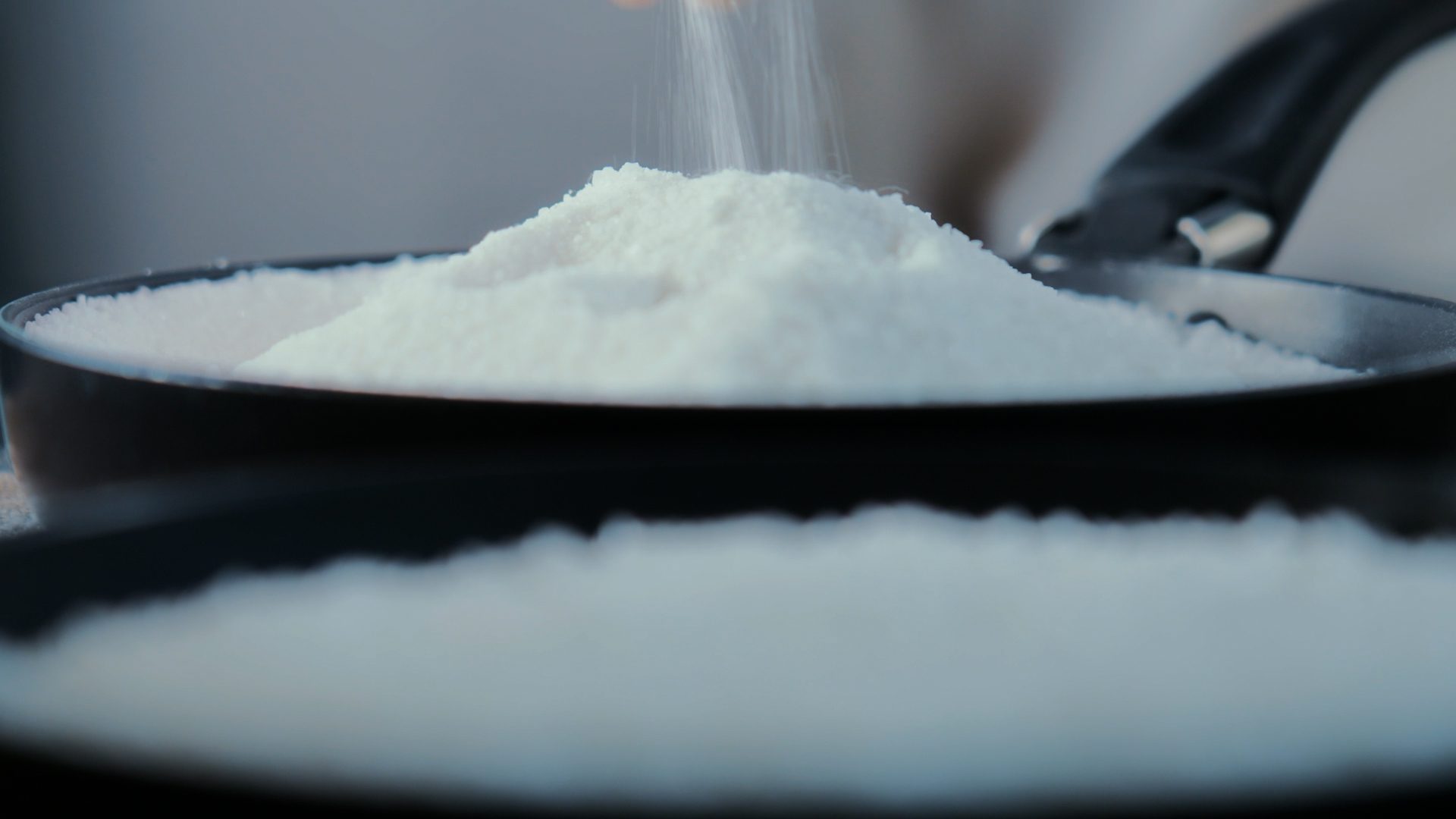 White powder, possibly flour or sugar, being poured onto a large pile in a black pan.