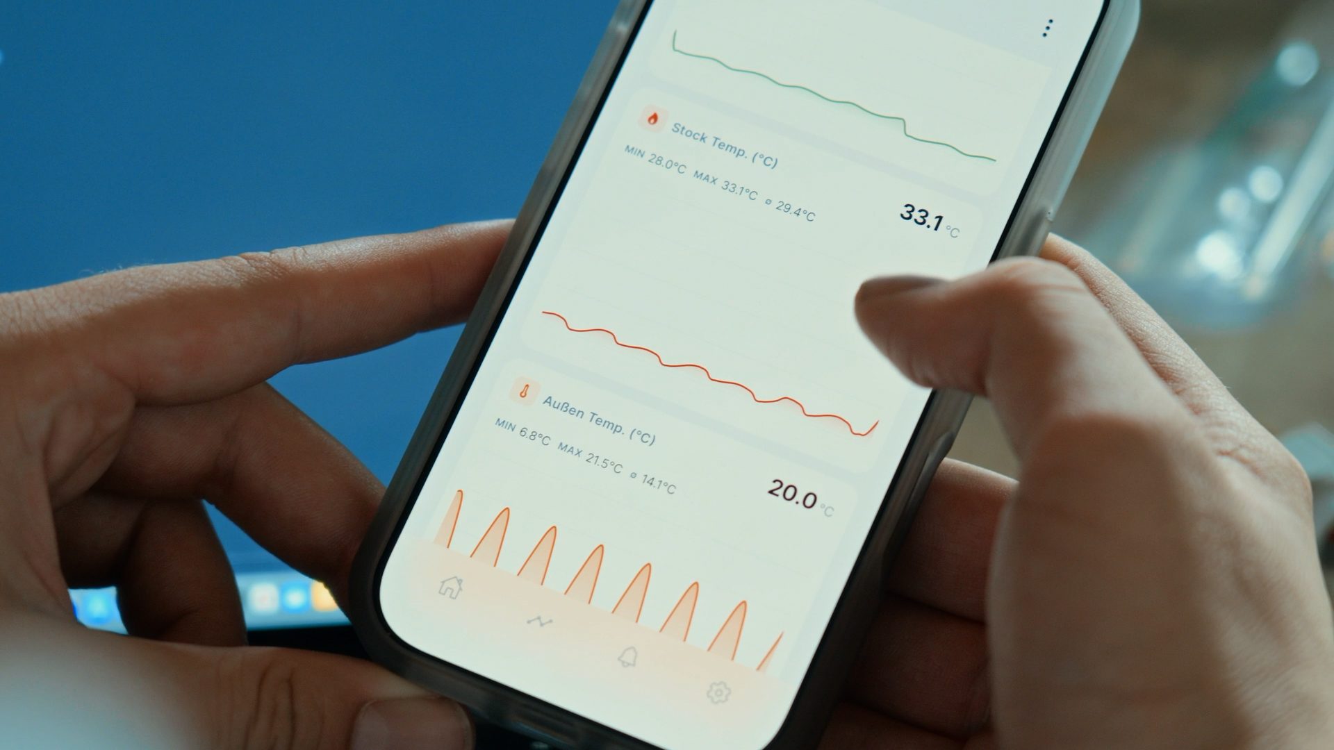 A person holds a smartphone displaying a temperature monitoring app with graphs and data for stock and outdoor temperatures.