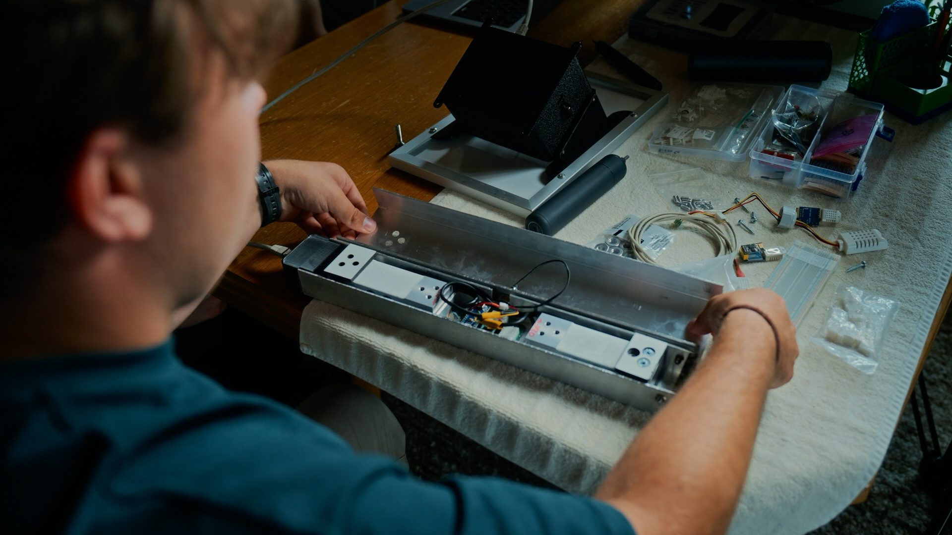 Person assembling electronics in a metal enclosure on a cluttered workbench with tools and components.