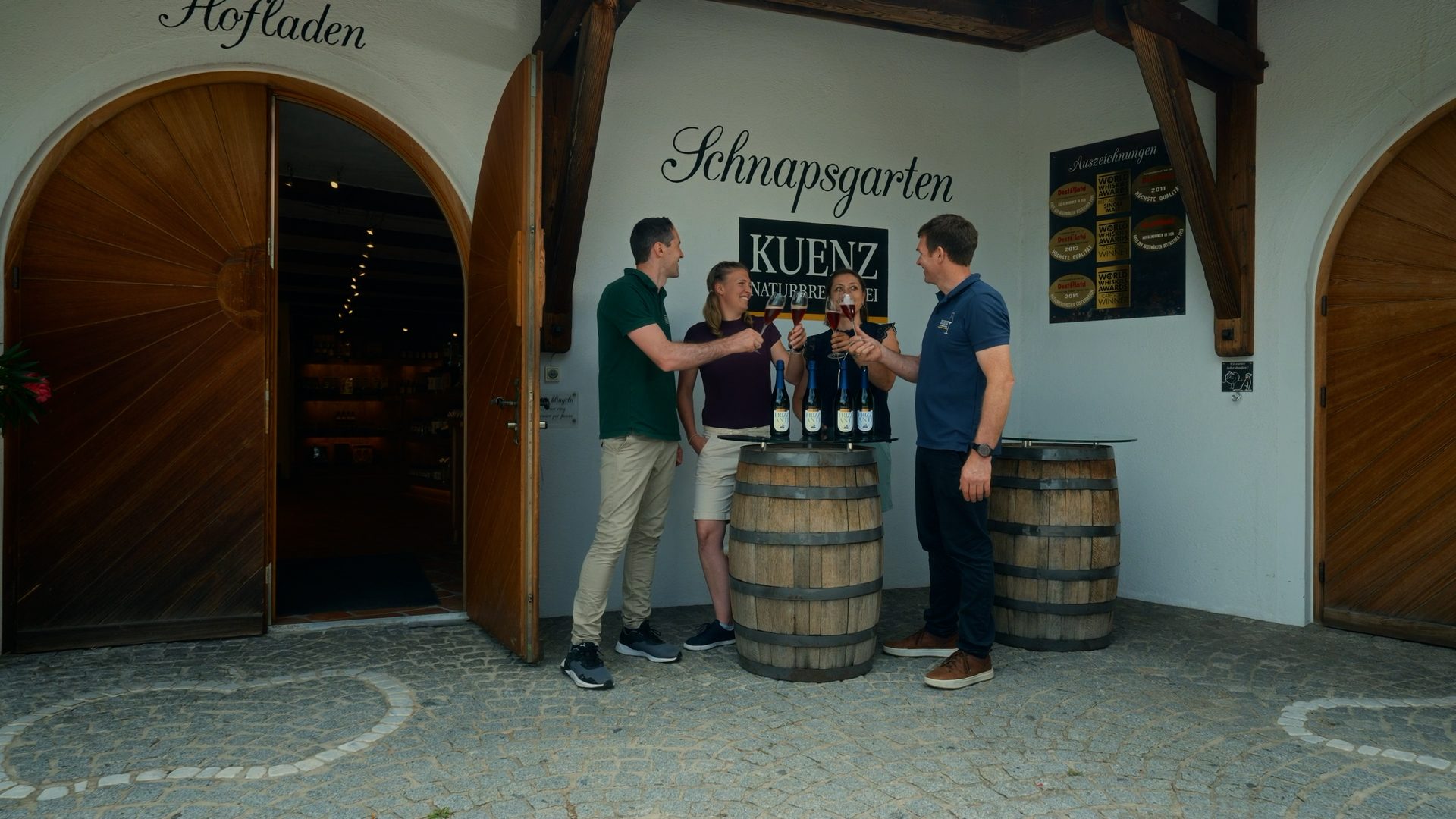 Four people toast with glasses outside the Schnapsgarten distillery, with barrels and bottles visible.
