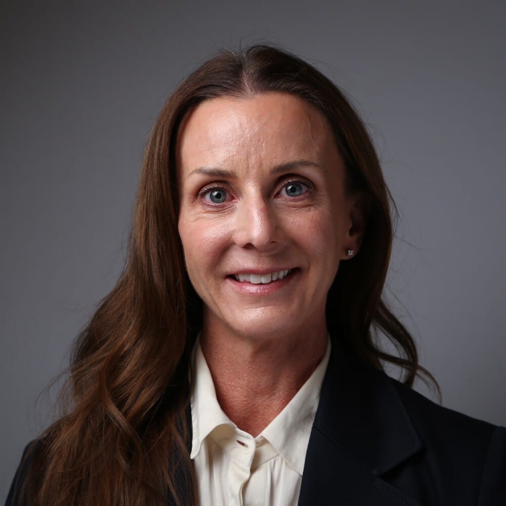 Smiling woman with long brown hair, blue eyes, wearing a blazer.