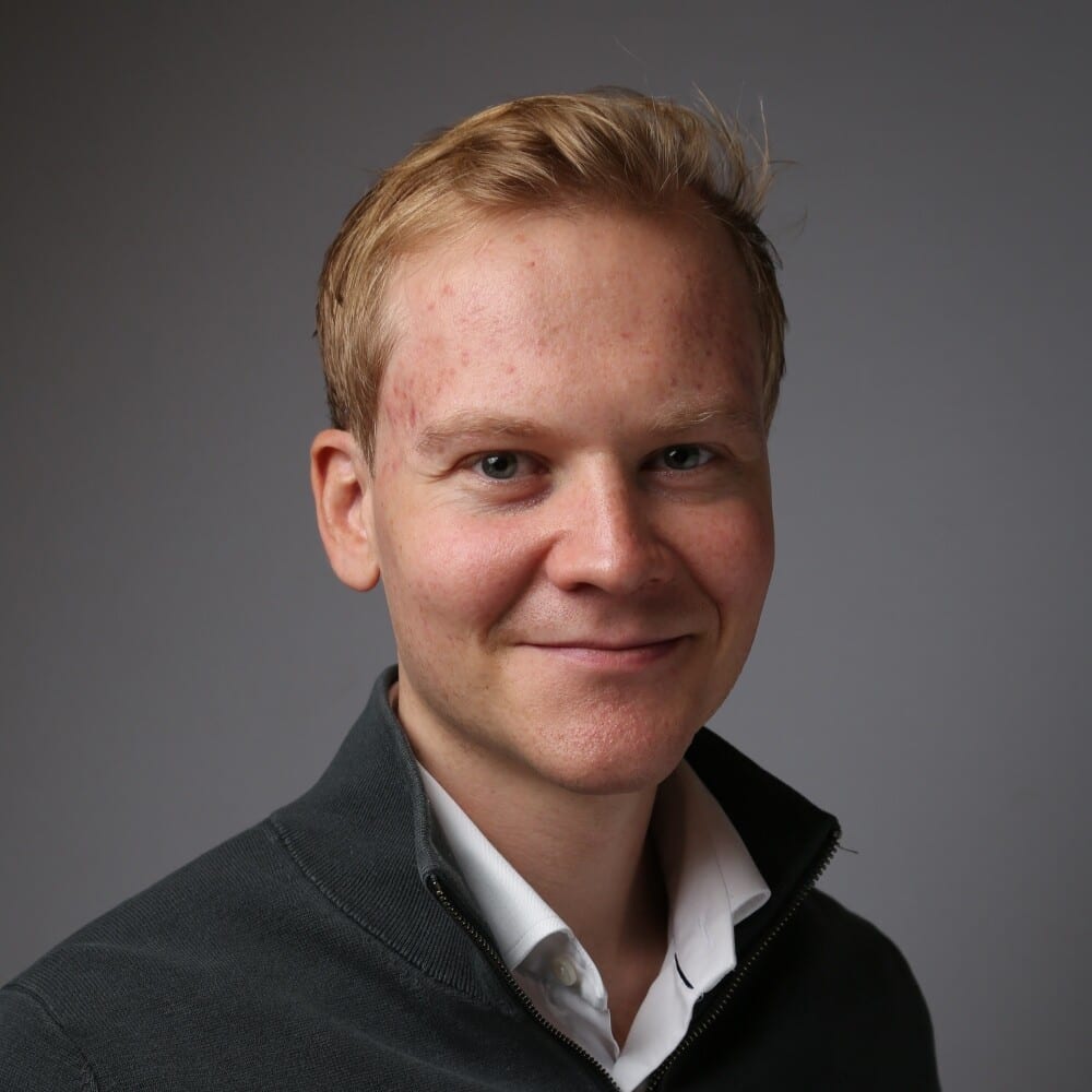 Young man with blonde hair, blue eyes, smiling in a gray sweater.