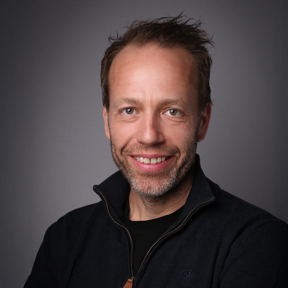 Smiling man with brown hair and beard in dark sweater, gray background.