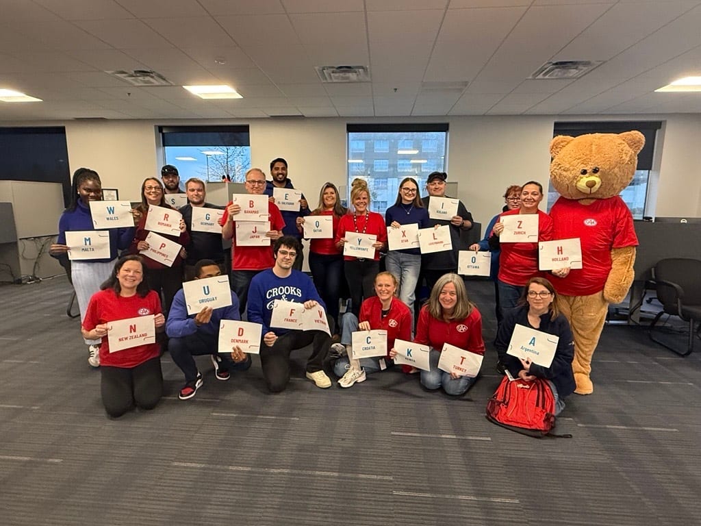 A group of people and a large teddy bear mascot in an office holding signs with letters and country names.