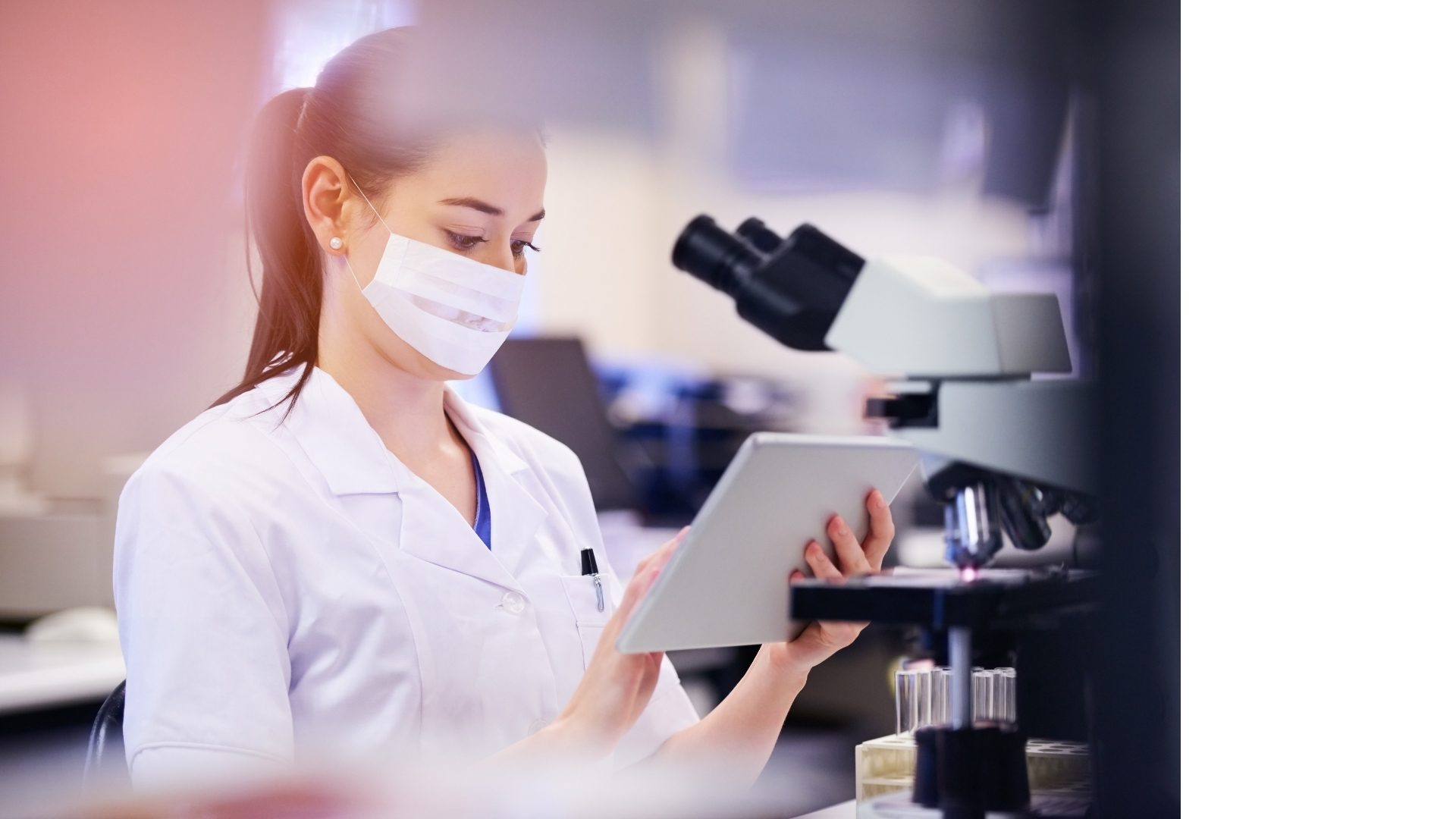 Female scientist in a lab coat and mask uses a tablet next to a microscope in a laboratory.