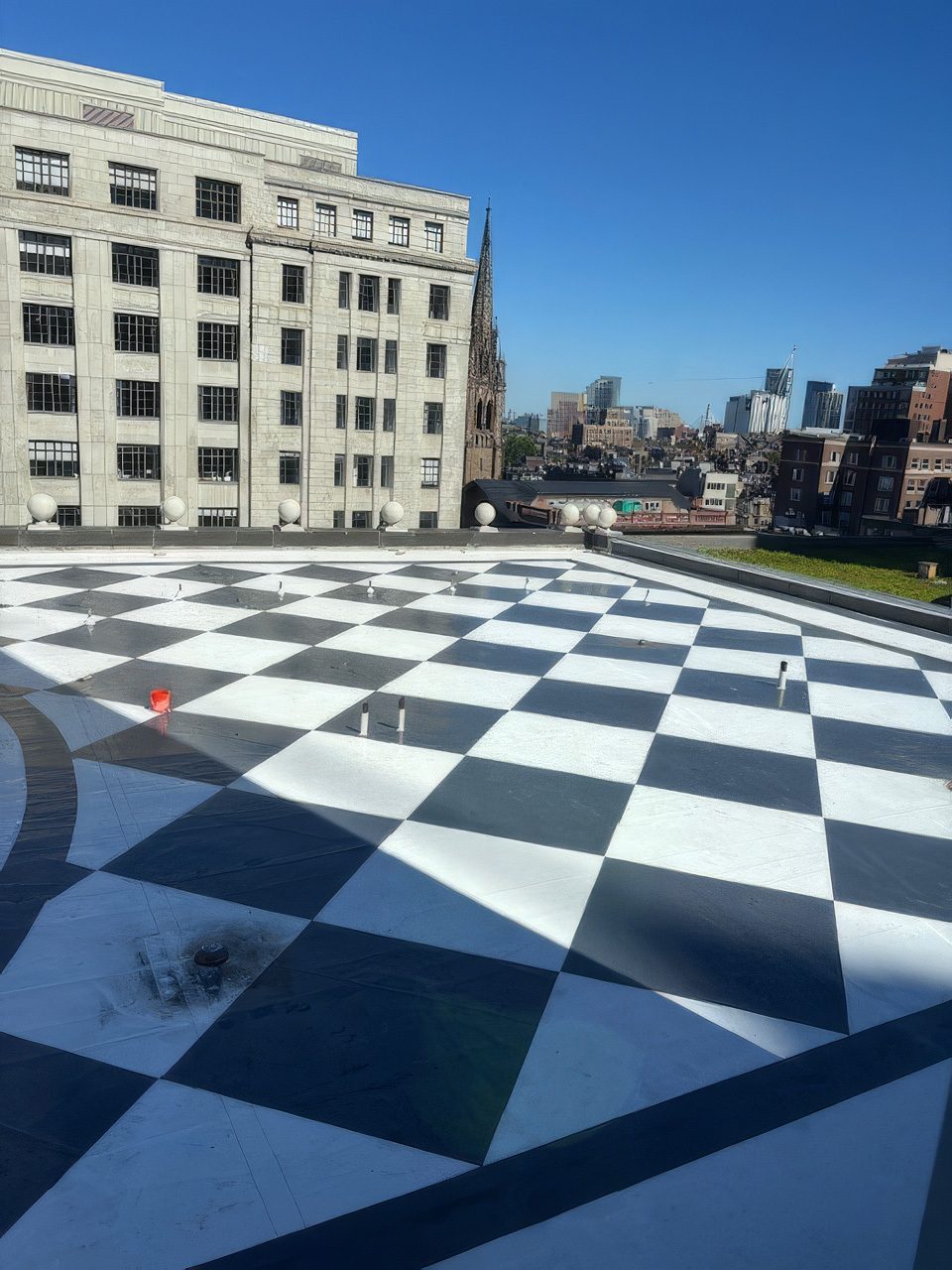 A checkerboard-patterned rooftop with a large building, church spire, and city skyline under a clear sky.