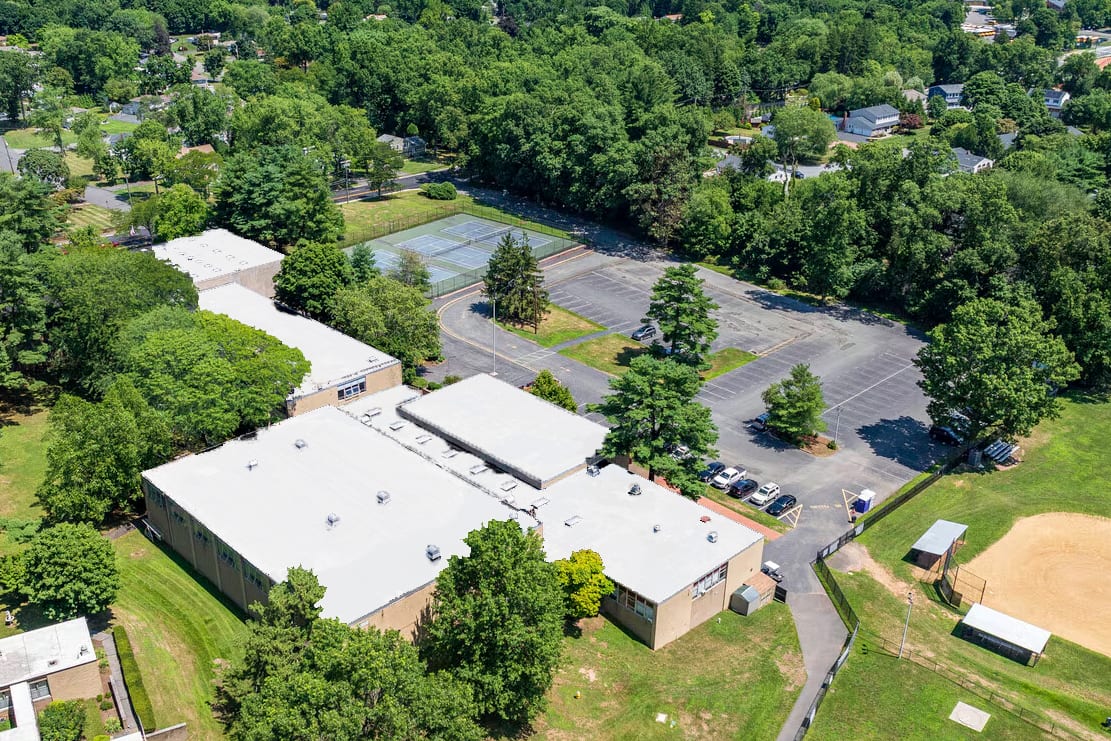 Aerial view of school buildings, tennis courts, baseball field, and parking, nestled among green trees.