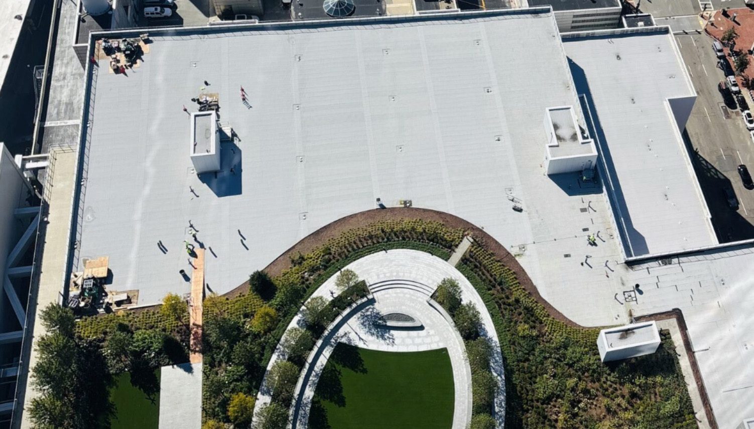 Aerial view: large building roof with workers on gray section and a circular green roof garden.