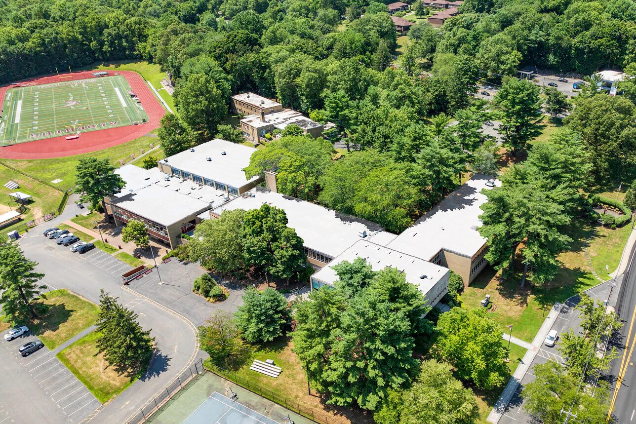 Aerial view of a school campus, including buildings, a football field, and dense surrounding greenery.
