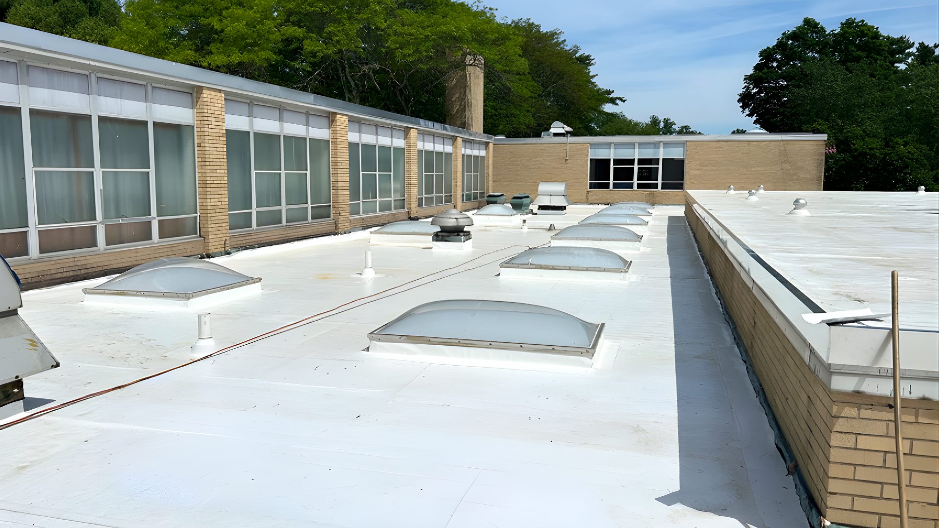 A flat white roof with several dome skylights, ventilation units, and a brick school building.