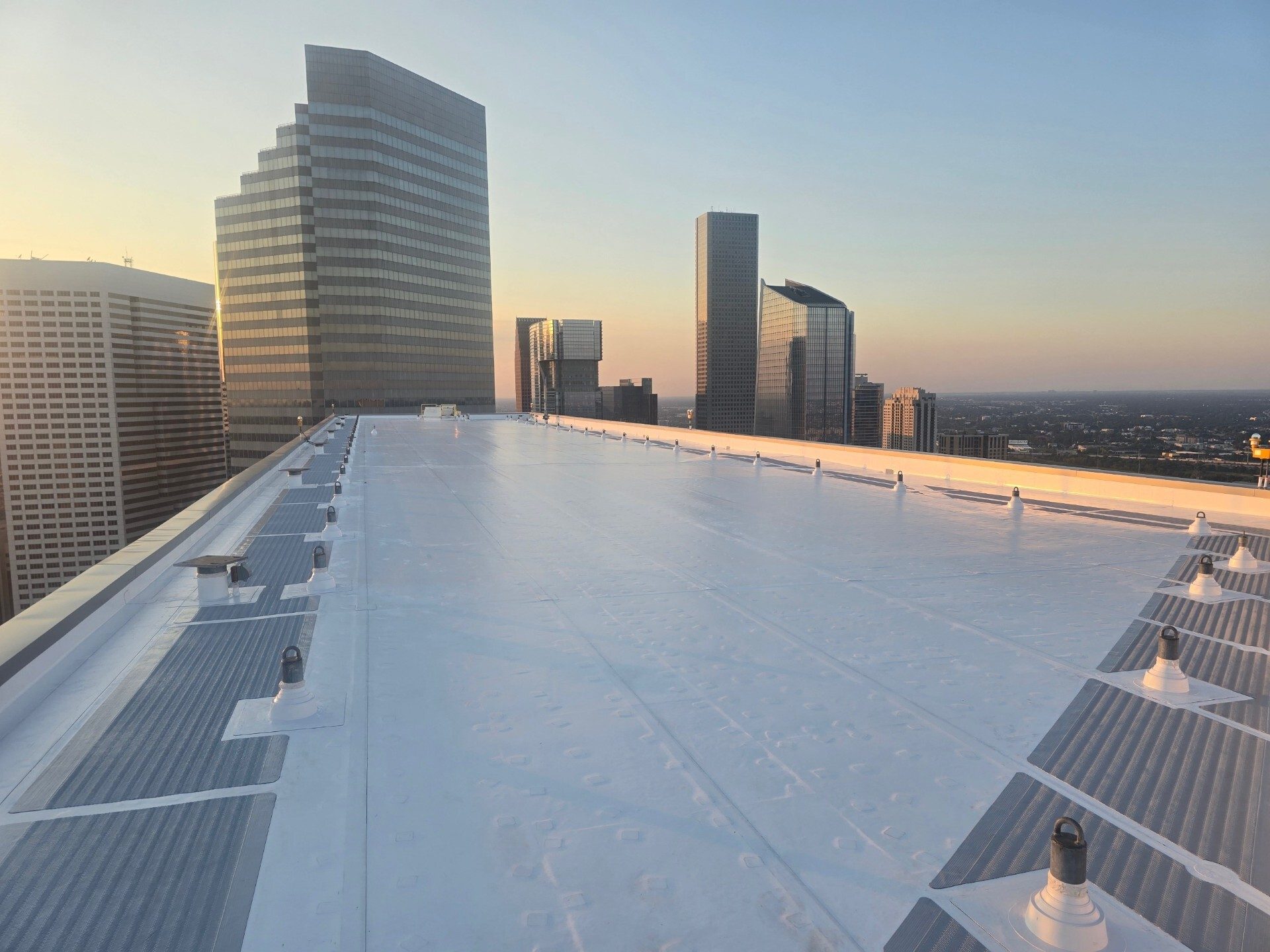 Rooftop view of a city skyline at sunset, with a white roof in the foreground and tall buildings.