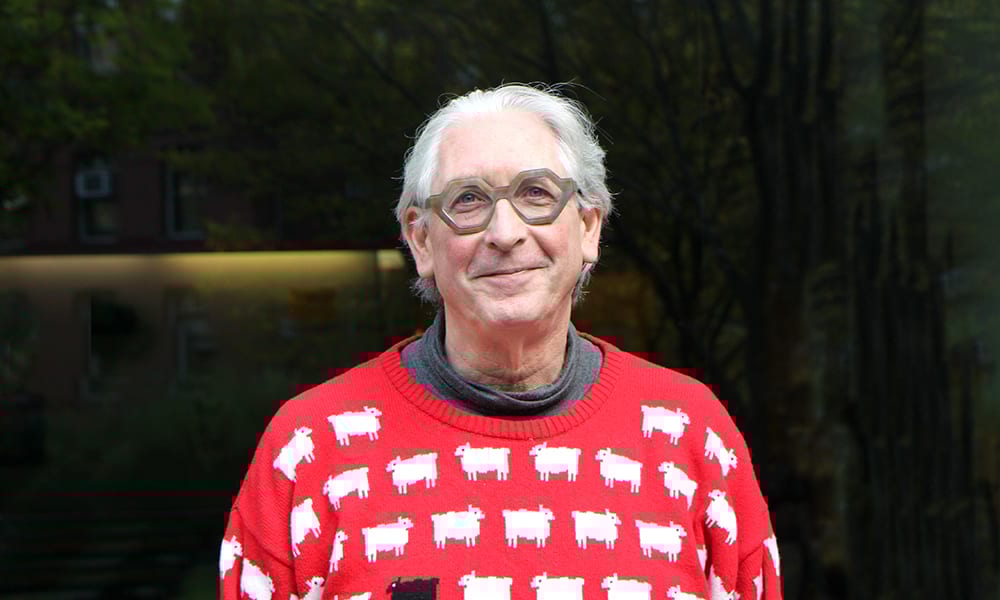 Smiling man with grey hair, unique glasses, in a red sweater with white and one black sheep.