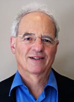 A smiling older man with glasses and gray hair, wearing a blue shirt and black jacket.