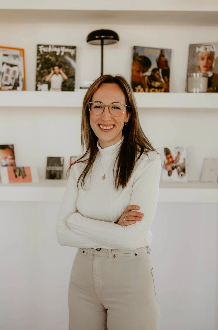 A smiling woman in glasses and a white turtleneck, arms crossed, standing before shelves.