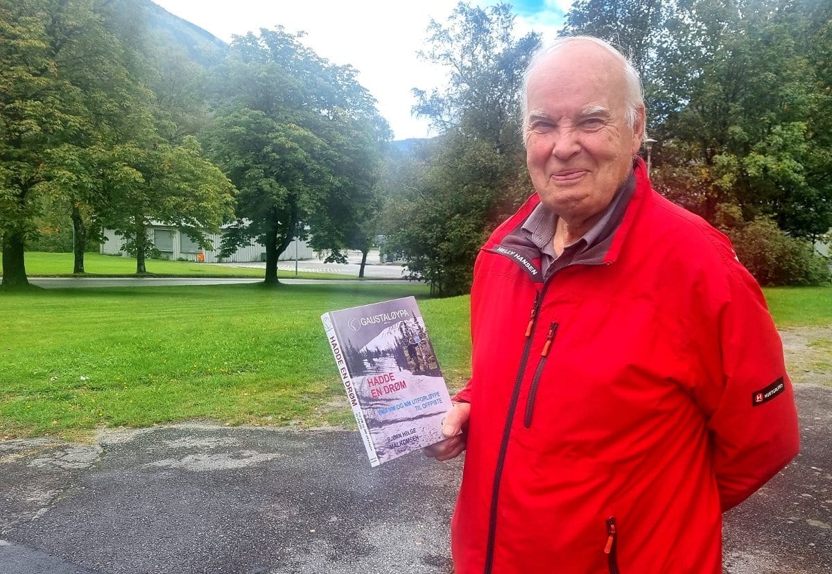 Older man in red Helly Hansen jacket holds "GAUSTALØYPA HADDE EN DRØM" book outdoors.