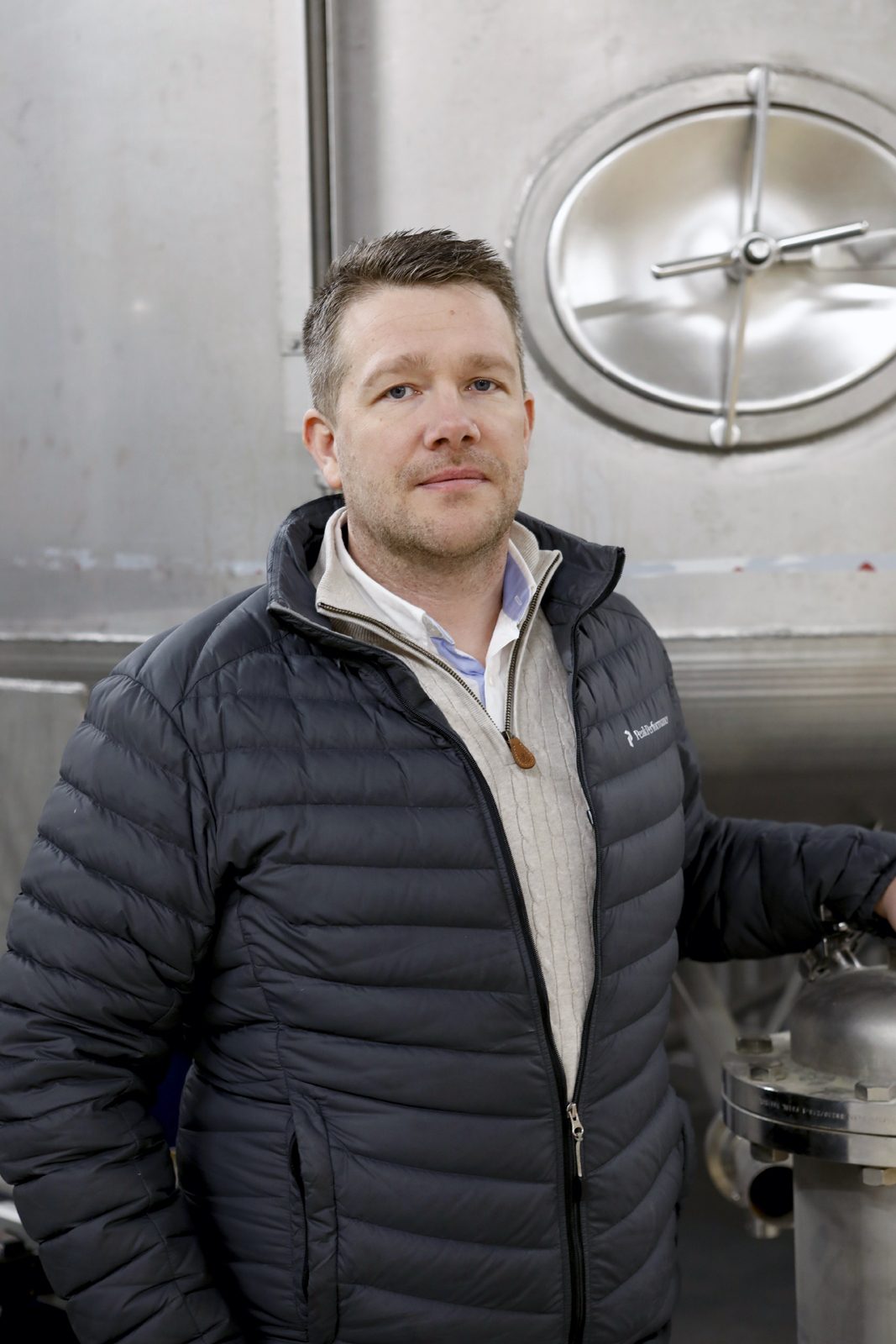 Man in black puffer jacket and sweater standing by a large metal tank with a circular hatch.