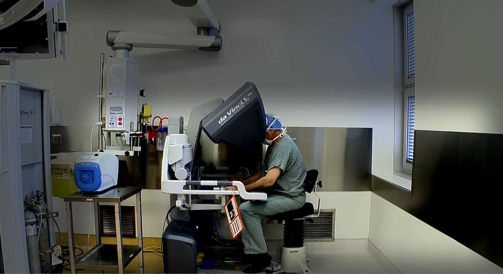 Surgeon operating a da Vinci surgical robot console in a modern operating room with various medical equipment.
