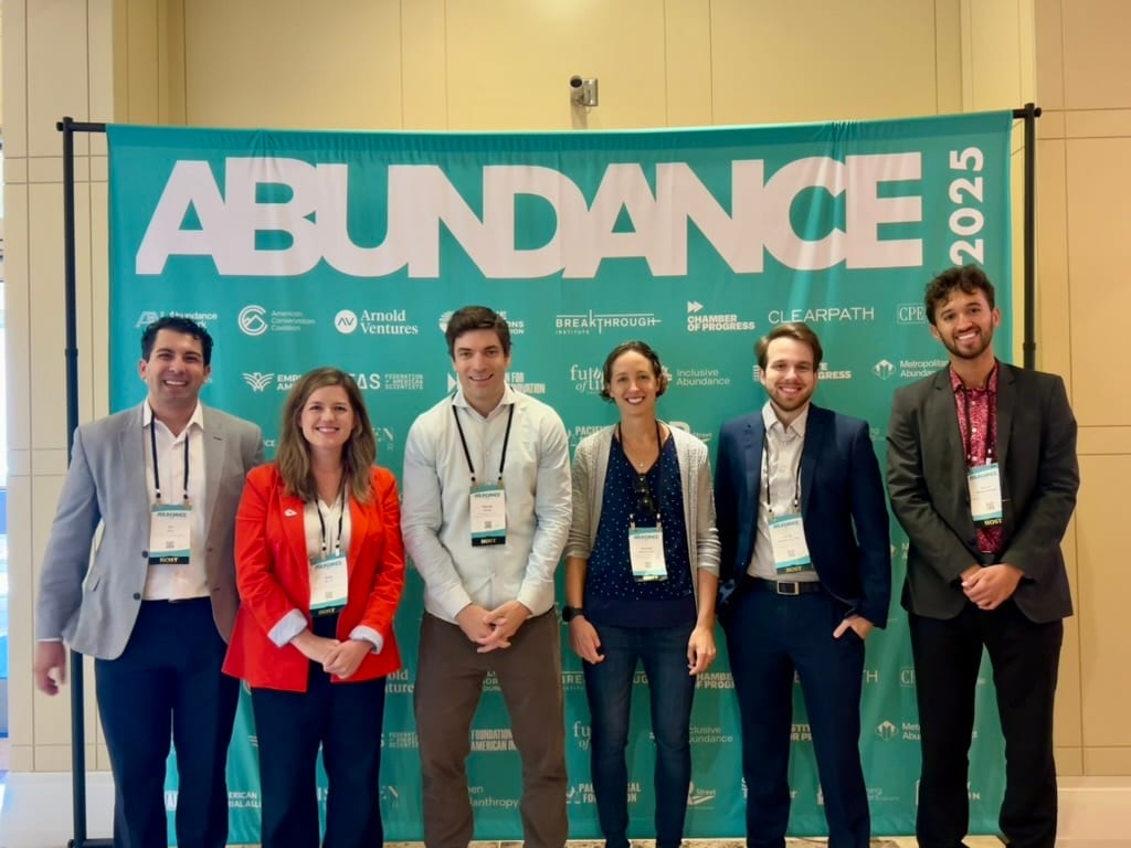Six people, three men and three women, stand in front of a teal banner displaying "ABUNDANCE 2025."