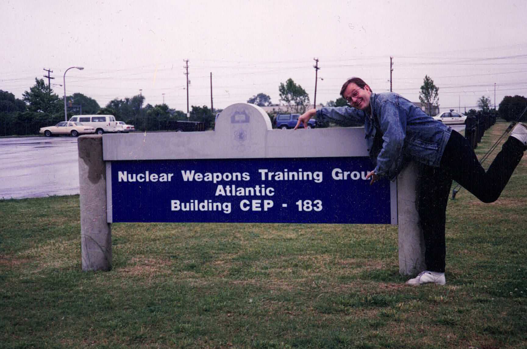Man playfully posing by a "Nuclear Weapons Training Group Atlantic Building CEP - 183" sign.