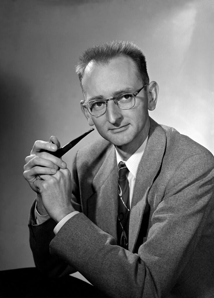 B&W portrait of a man in glasses and suit, holding a pipe, looking at the camera.