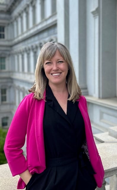 Smiling woman, blonde, wearing a bright pink blazer, by a classical building.