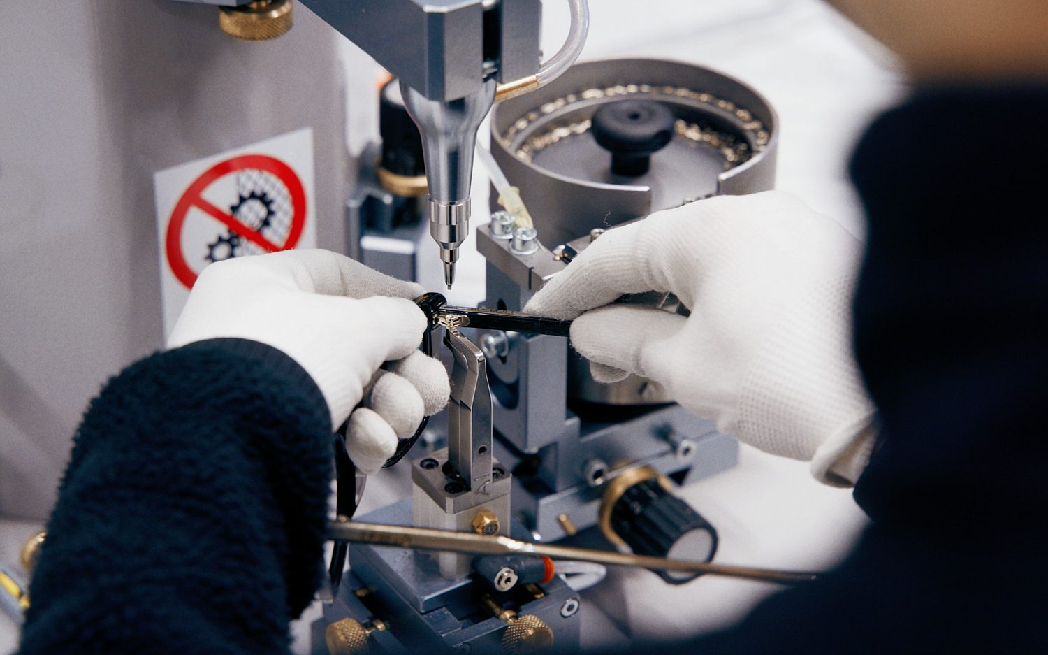 Close-up of gloved hands assembling a small part with precision machinery, safety sticker visible.