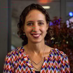 Smiling woman with dark curly hair and light eyes in a colorful patterned shirt.