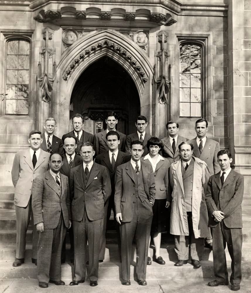 B&W group of formally dressed men and one woman on stone building steps.