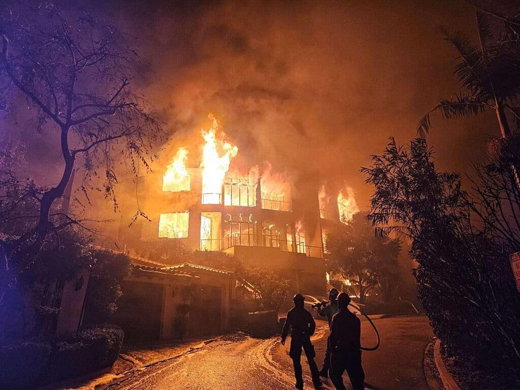 A large house is engulfed in bright orange flames at night, with three firefighters on the road below.