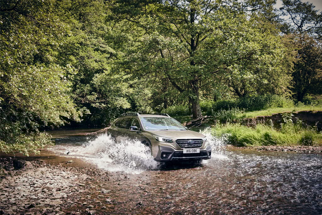 Khaki Subaru Outback SUV splashing through a river in a forest.