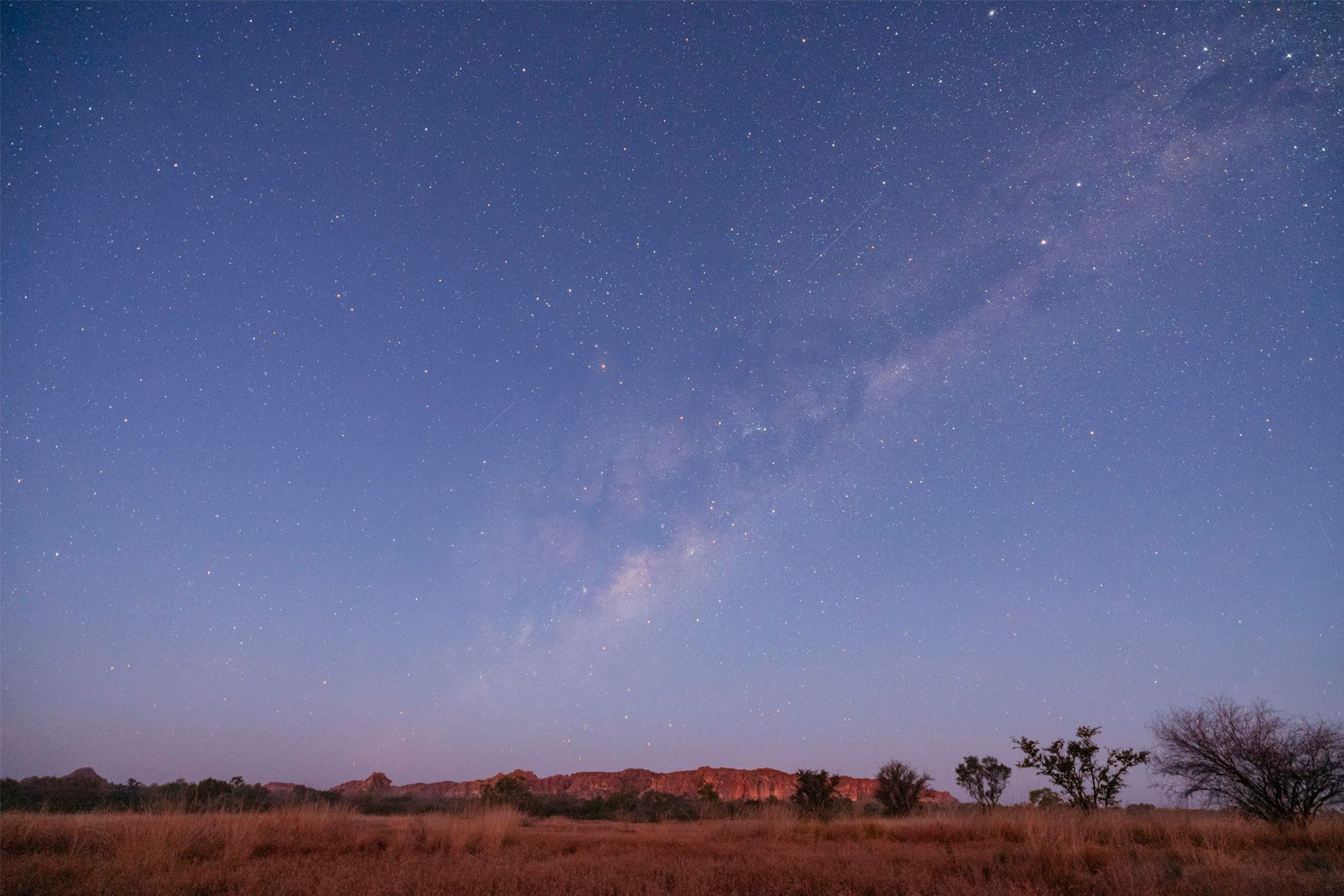 Milky Way streaking across a starry night sky above a reddish landscape with distant hills and trees.