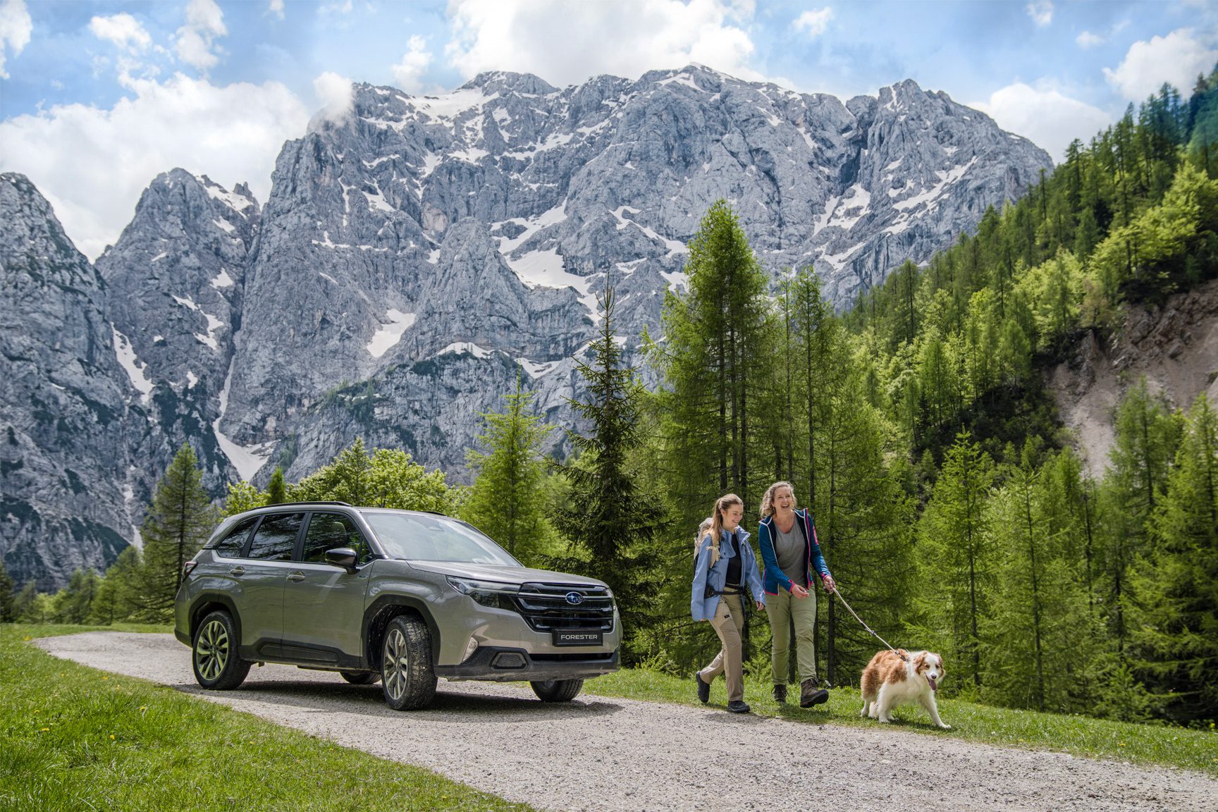Subaru Forester SUV on a mountain road with two women and a dog, snowy mountains and forest.
