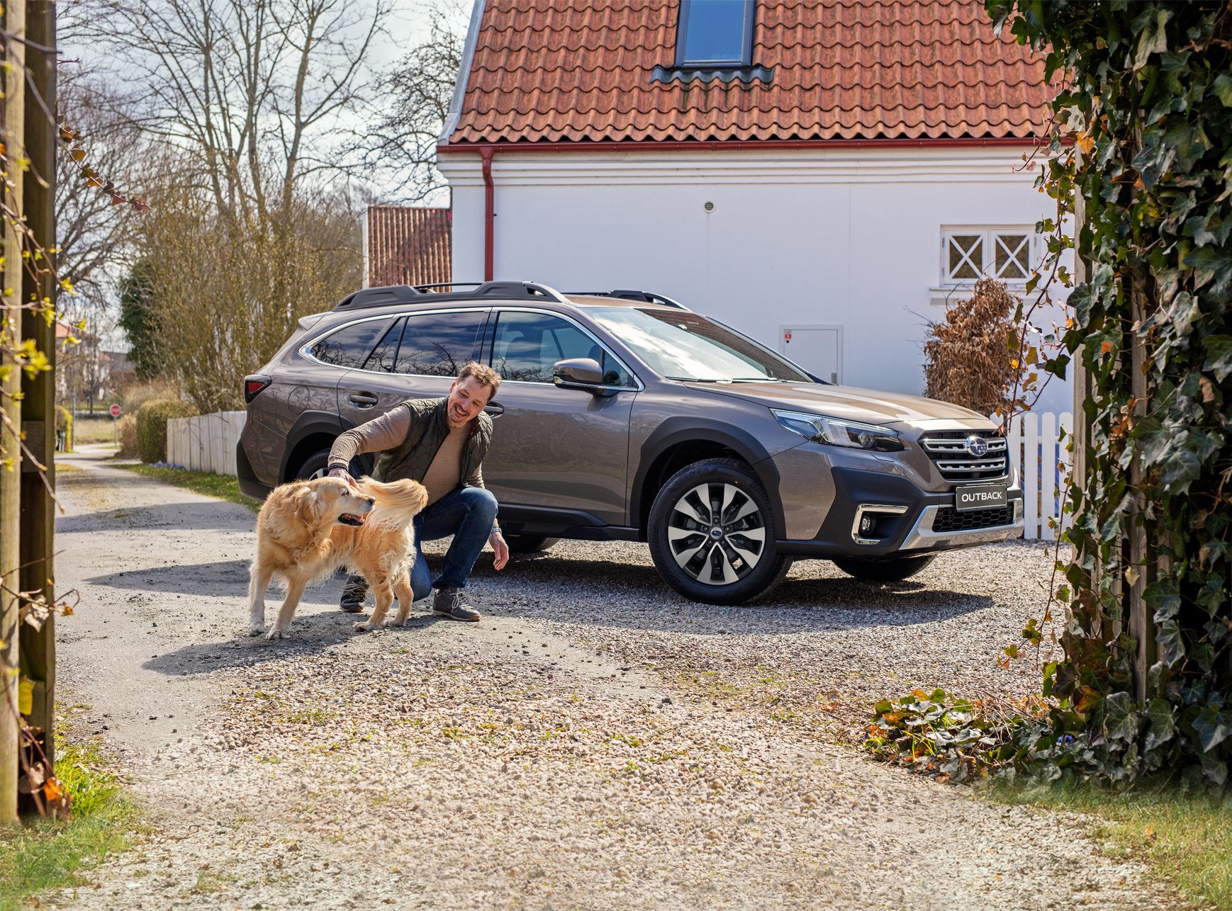 Man petting a golden retriever next to a gray Subaru Outback.