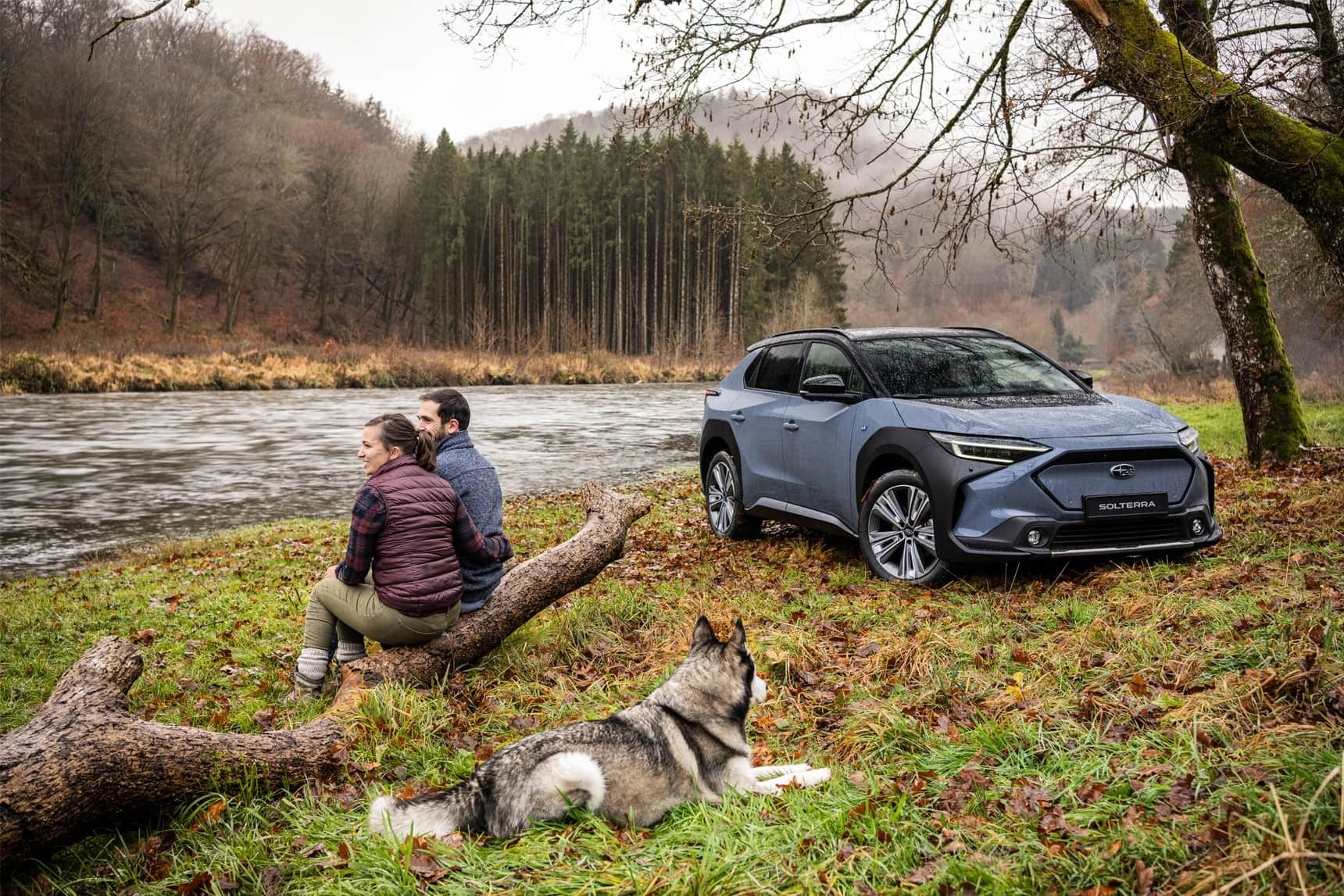 Couple and dog by a river with a blue Subaru Solterra SUV.